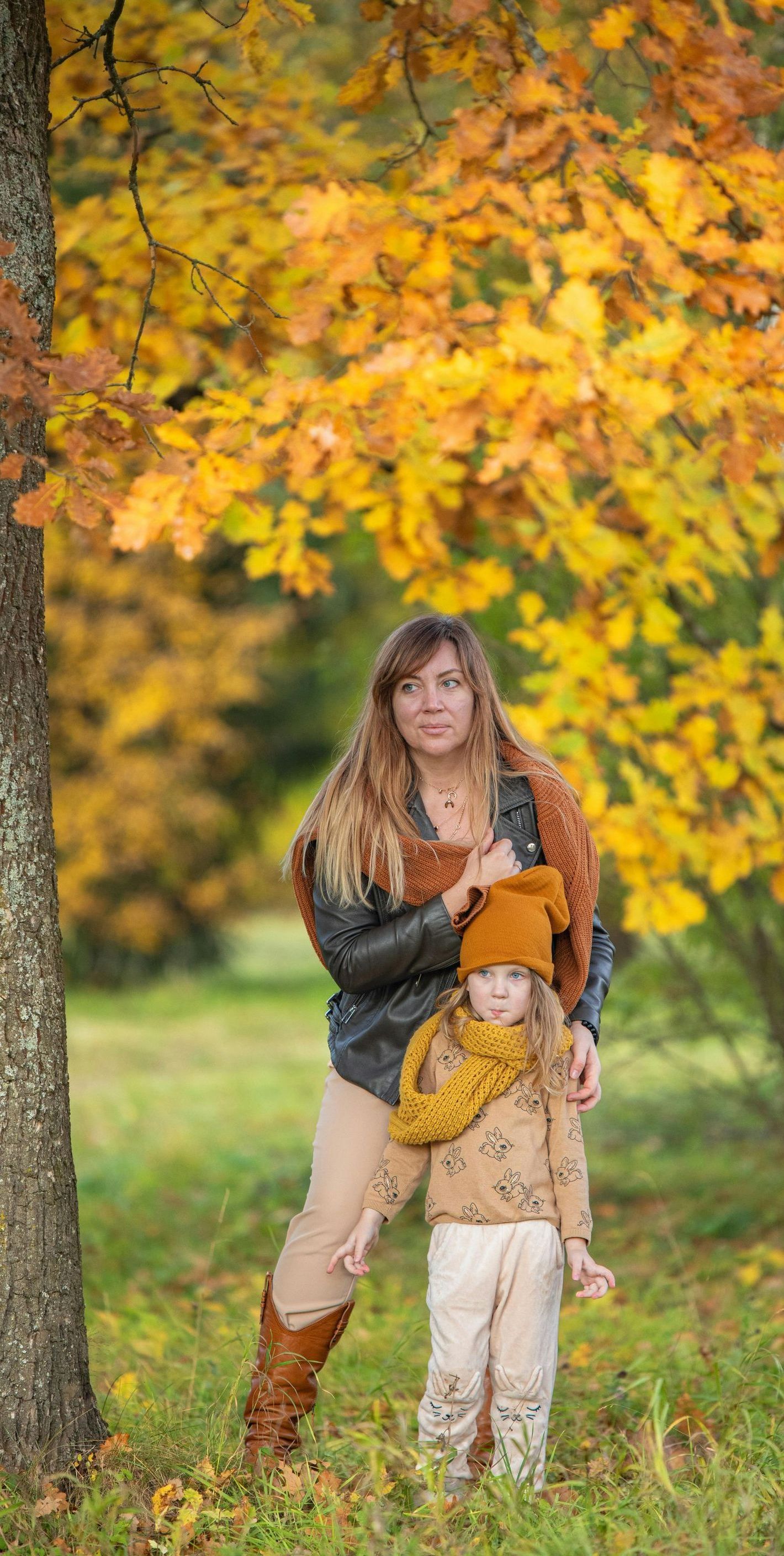 A woman and a little girl are standing next to a tree in a park.