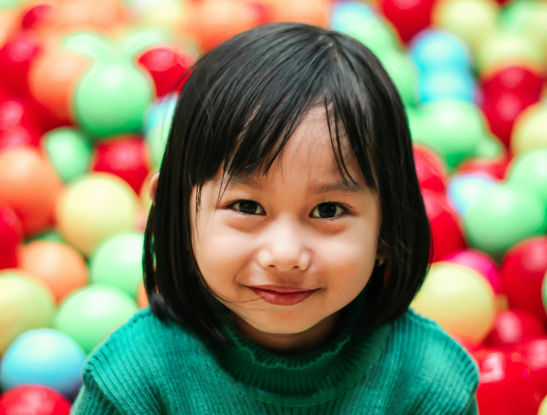 A little girl in a green sweater is sitting in a ball pit.