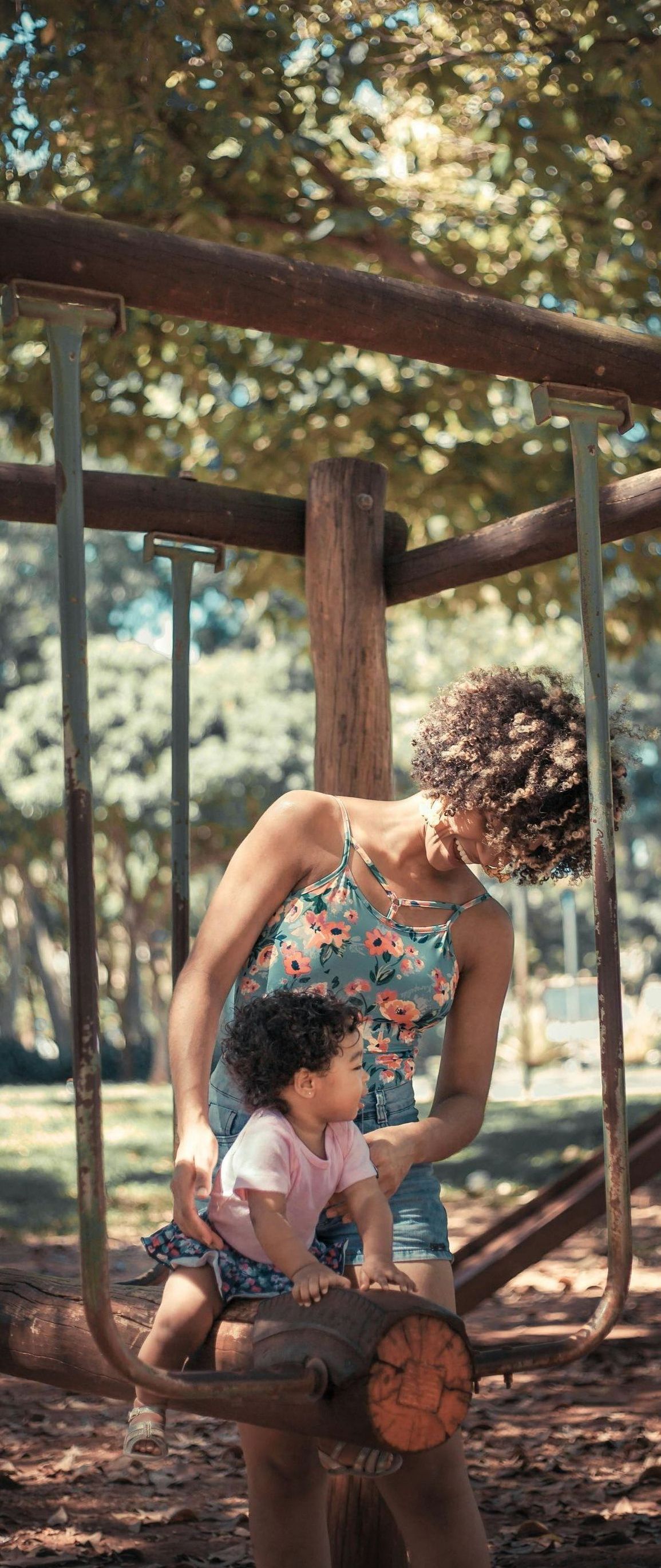 A woman and a child are playing on a wooden swing in a park.