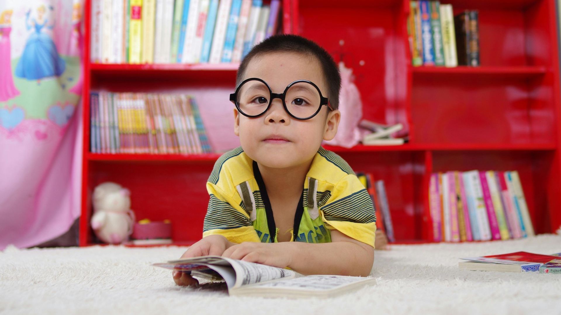 A young boy wearing glasses is laying on the floor reading a book.