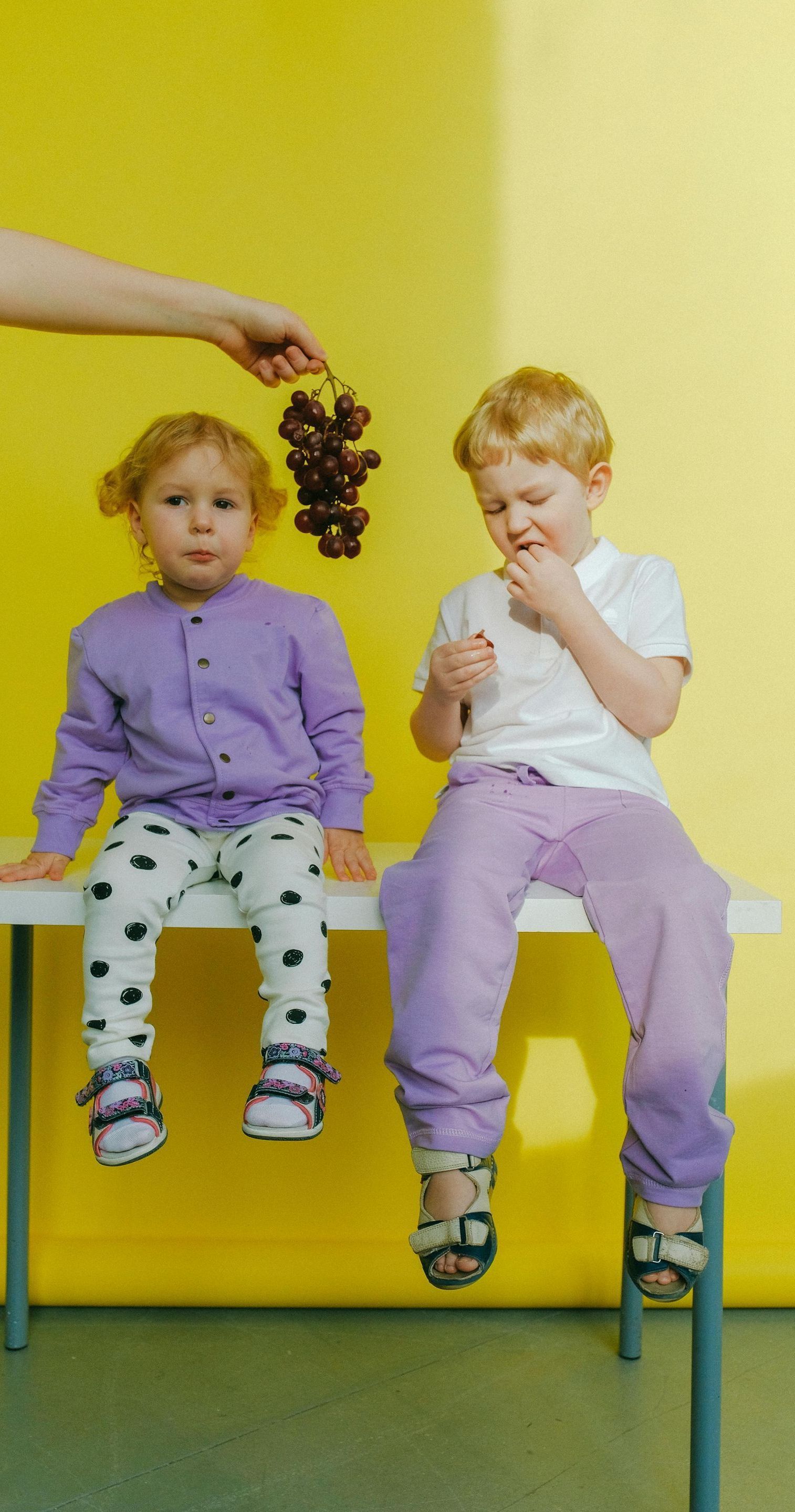 A boy and a girl are sitting on a table eating grapes.
