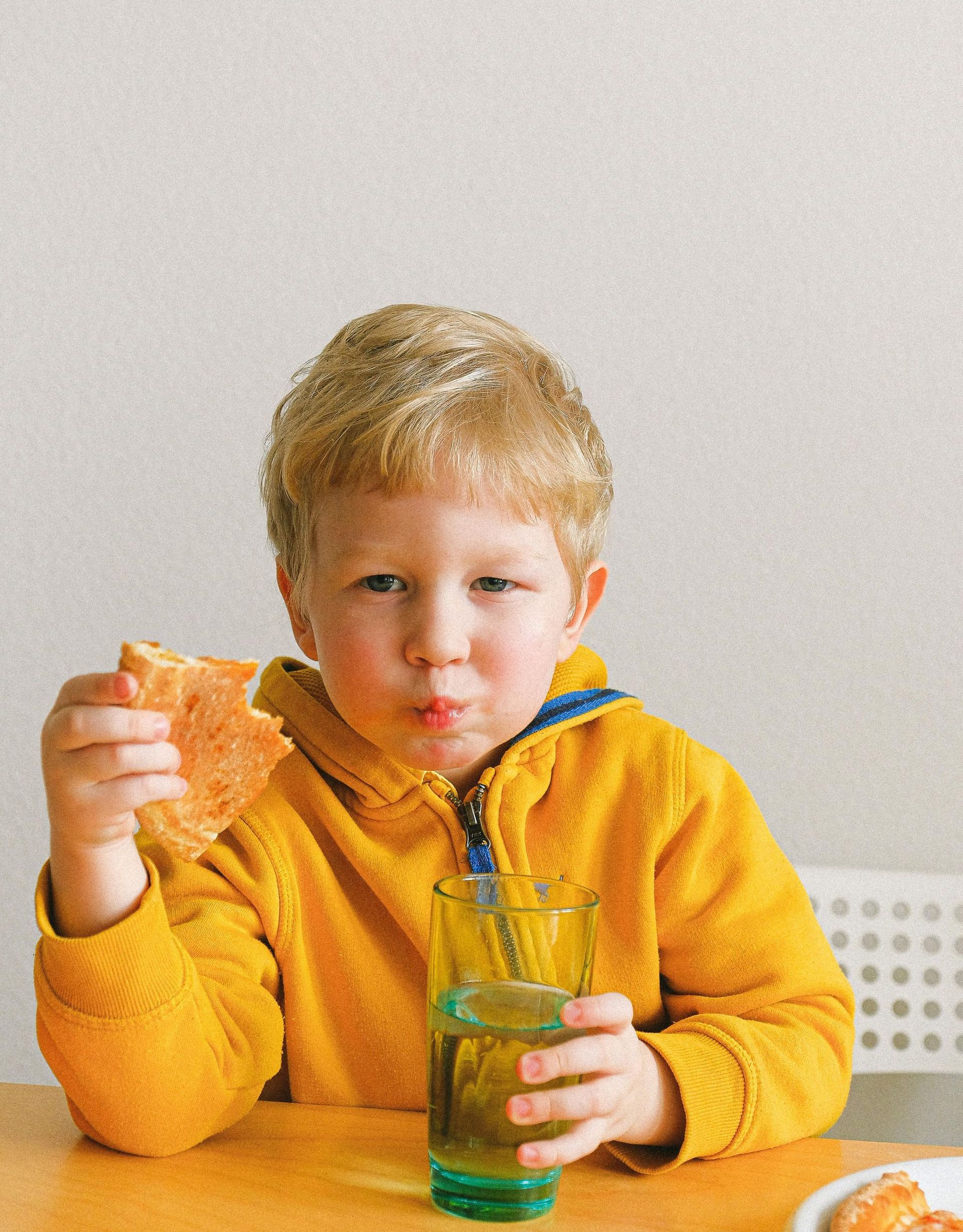 A young boy is sitting at a table eating a slice of pizza and drinking a glass of water.