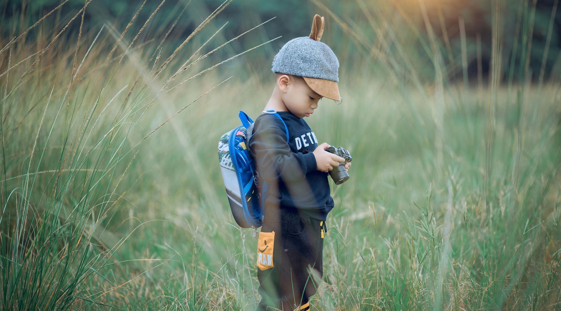A young boy with a backpack is standing in a field holding a camera.