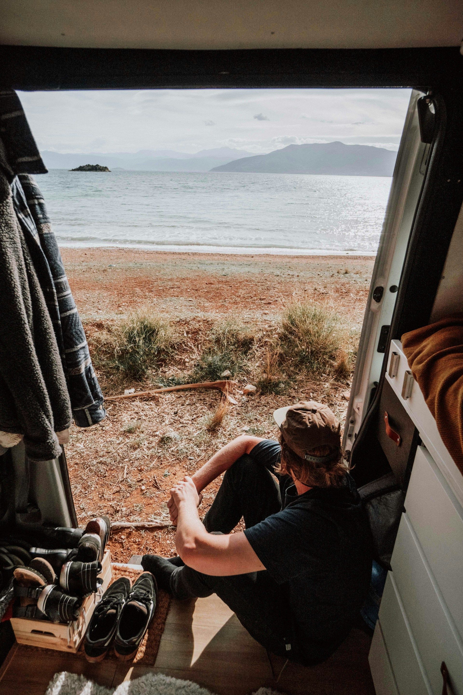 Person sitting in van doorway, looking at a beach and mountains. Ocean and shoreline visible.