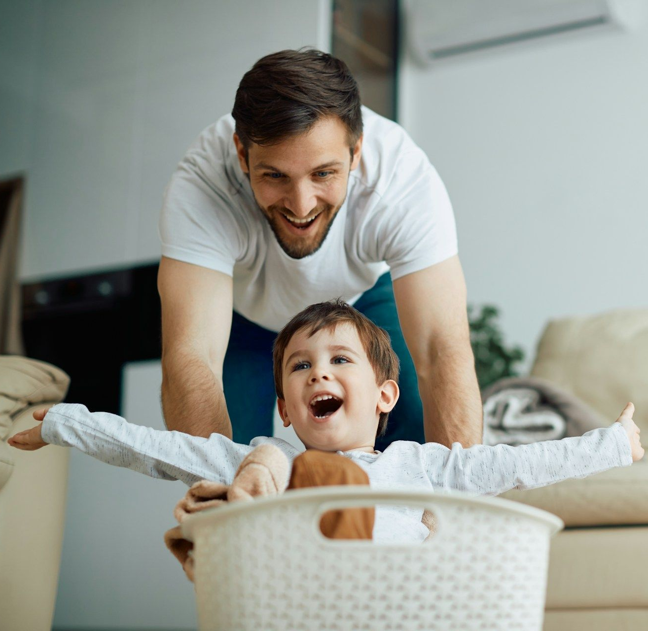 Man pushing a child in a laundry basket, both smiling and laughing indoors.
