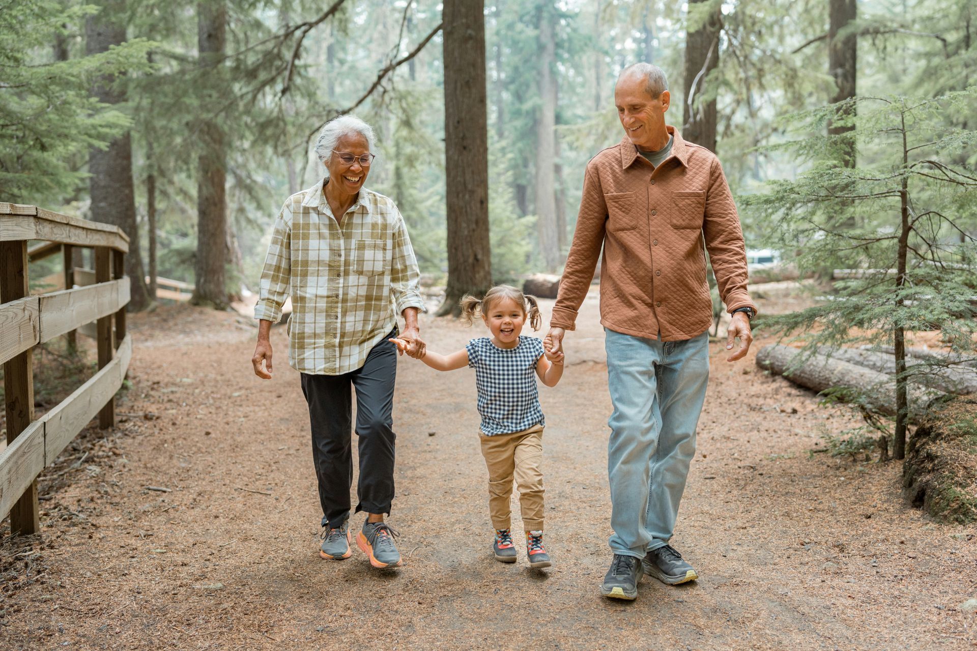 A young child holds hands with two adults while walking on a forest path.