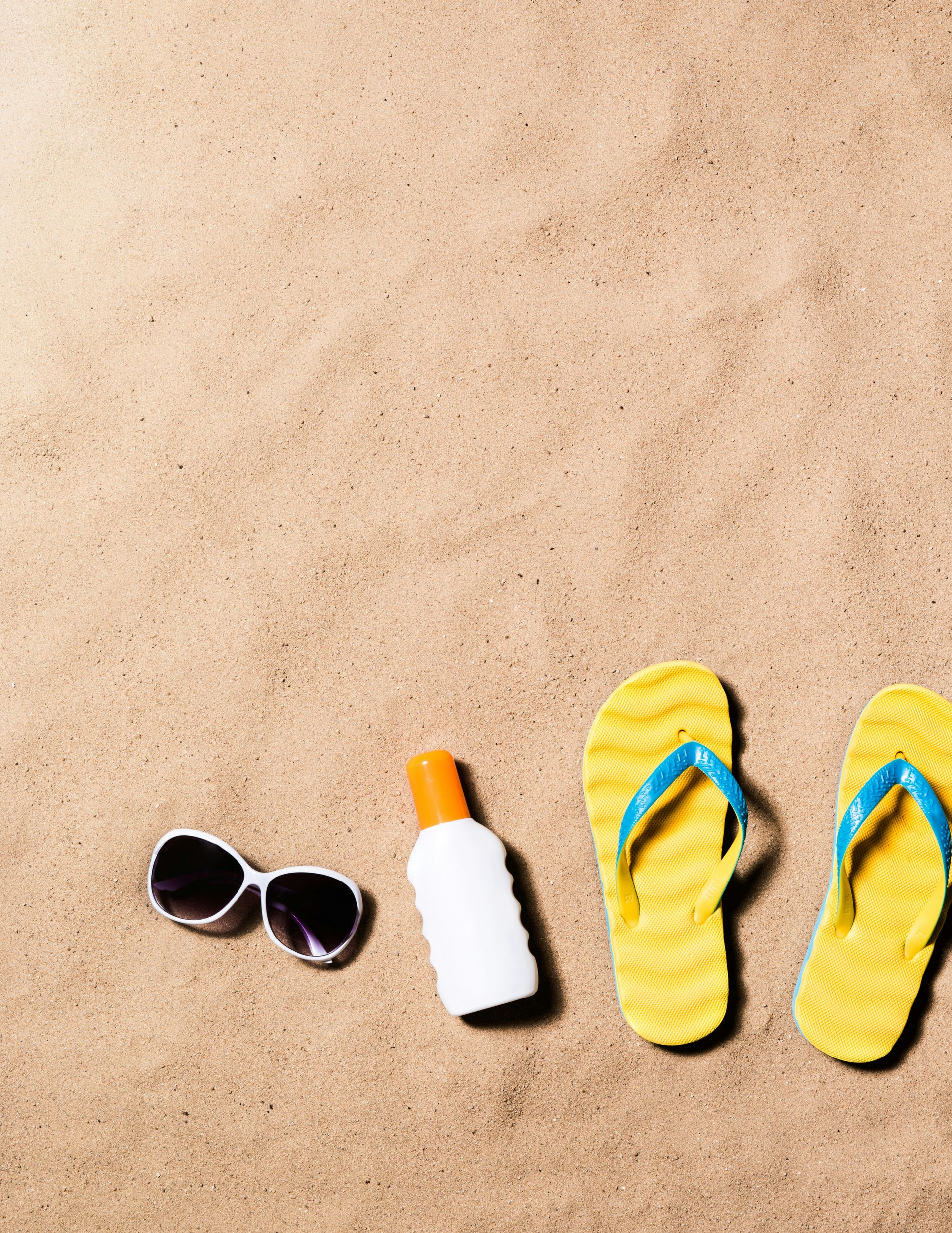 Sunglasses, sunscreen, and yellow flip-flops on a sandy beach.