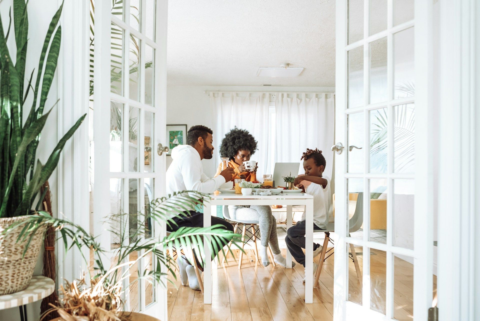 Family at a table, using a laptop and eating. The room has large windows and plants.