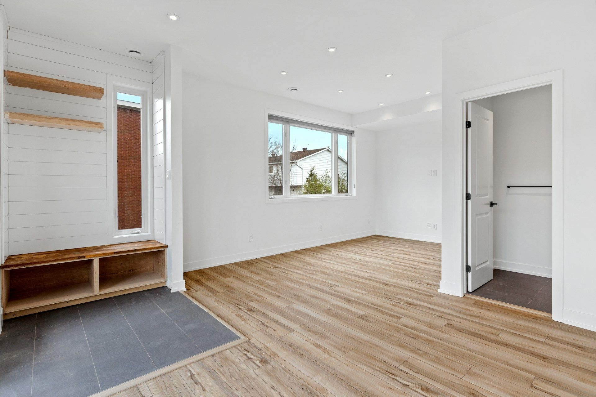 Empty, bright room with wood floors, white walls, built-in shelves, bench, and open door.