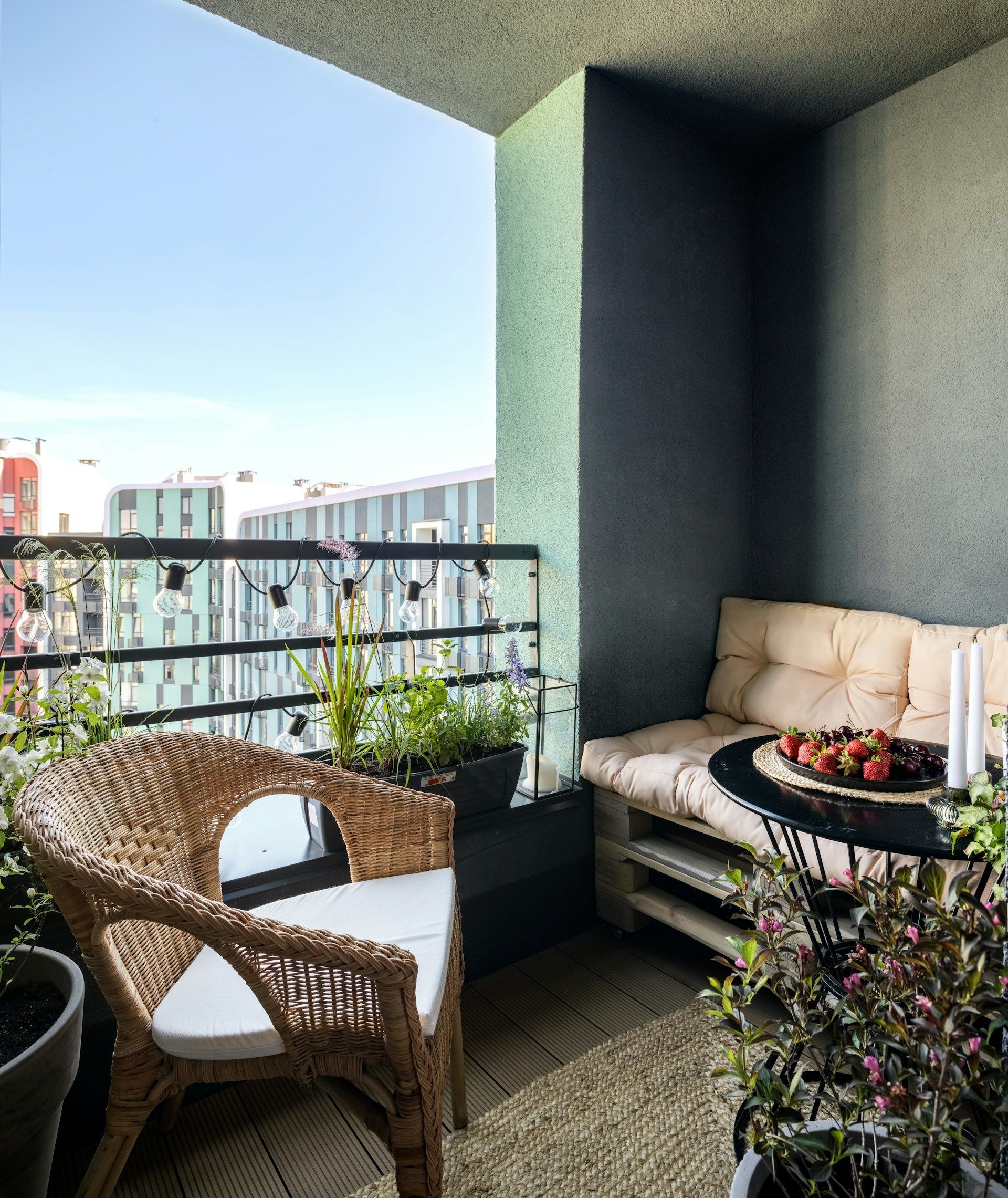 Cozy balcony with wicker chair, pallet sofa, small table with fruit, and plants. Black railing and blue sky.