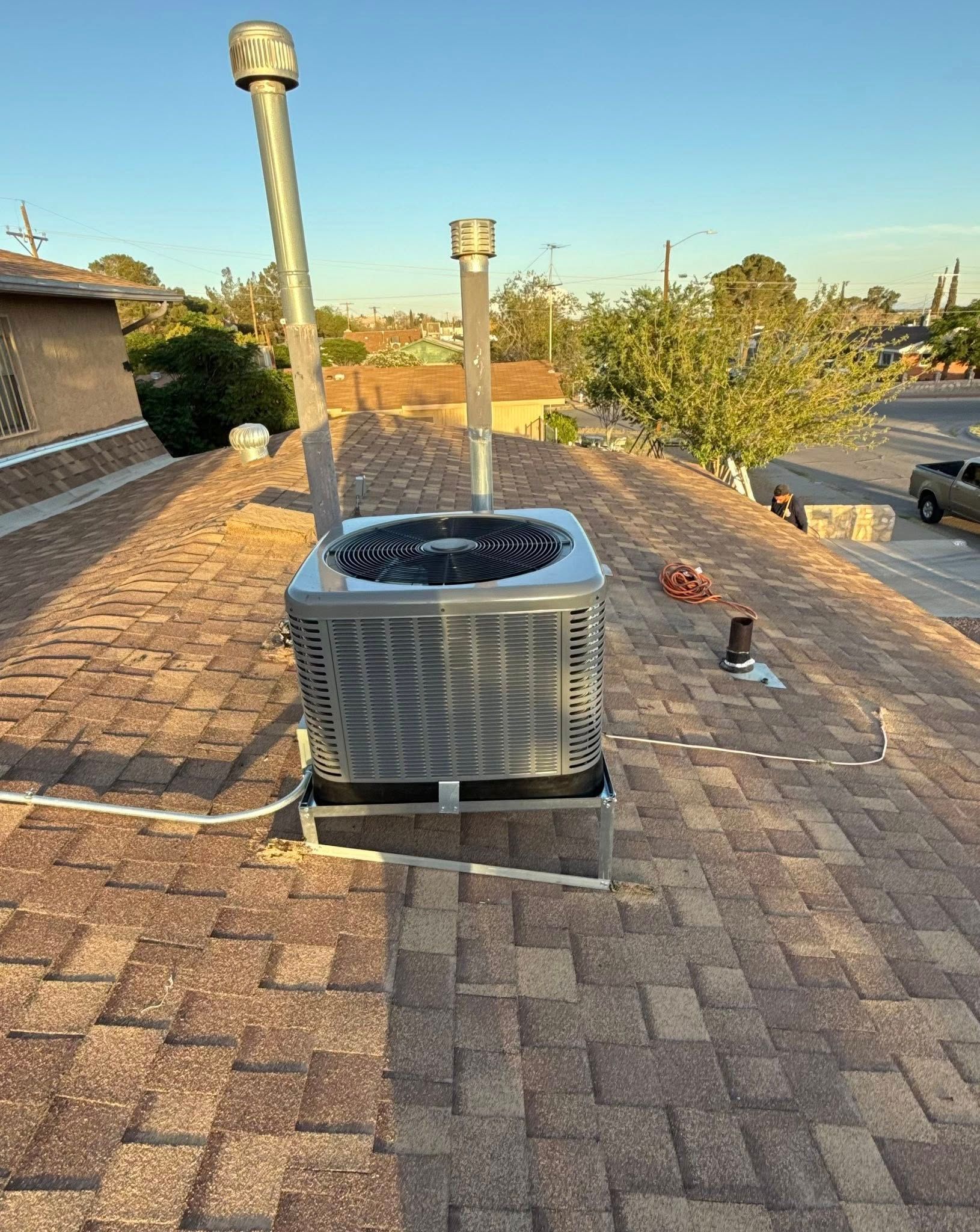 Air conditioning unit on a brown shingle roof with two vertical metal pipes; bright sky in background.