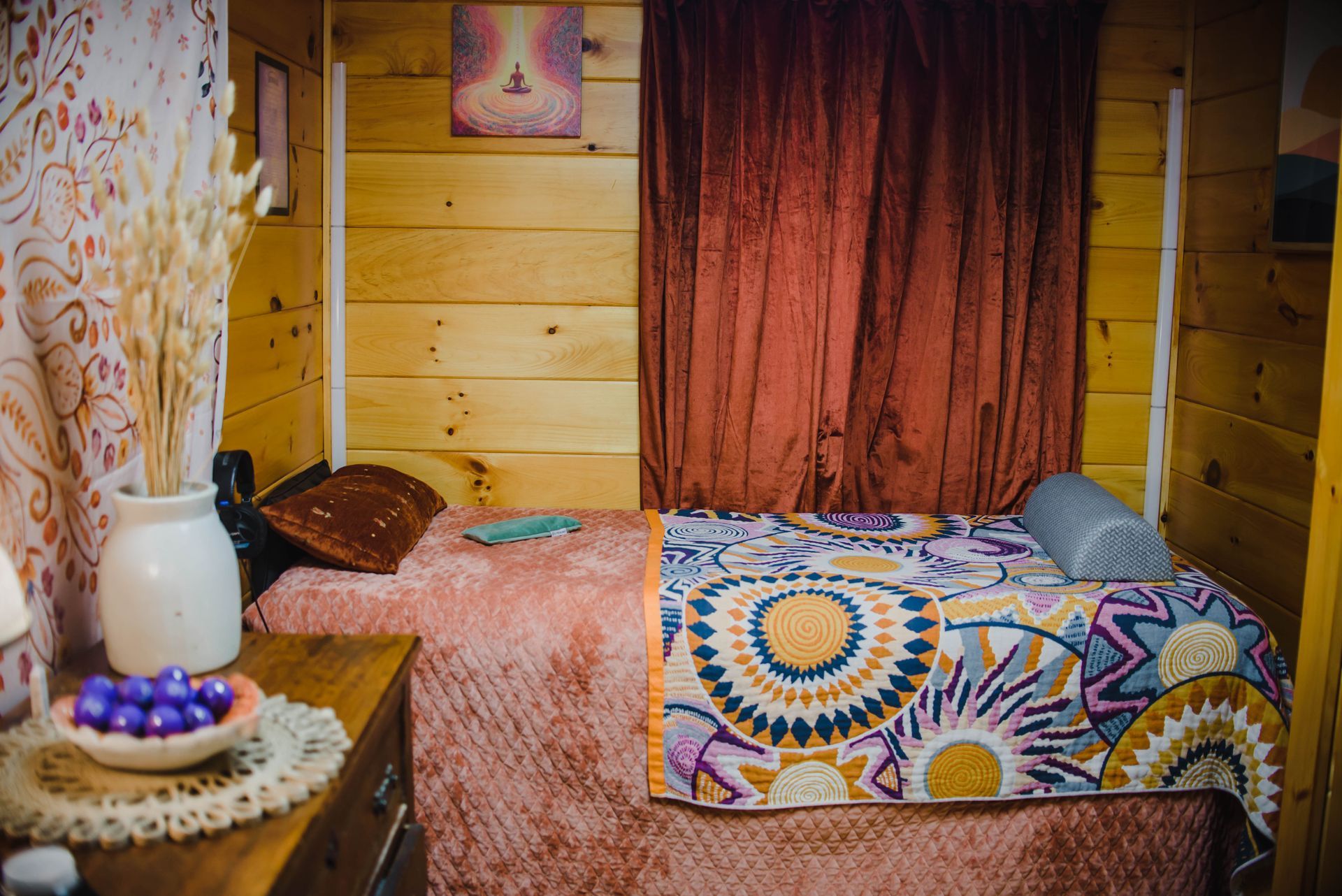 Cozy wooden bedroom with a single bed, colorful quilt, burgundy curtains, and a small table with vase and fruit.