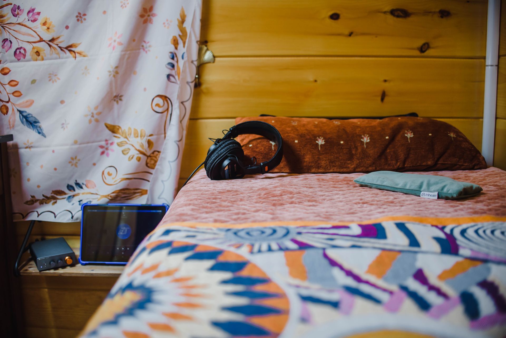 Bed with colorful quilt, headphones on pillow, and a laptop beside a wooden wall and curtain