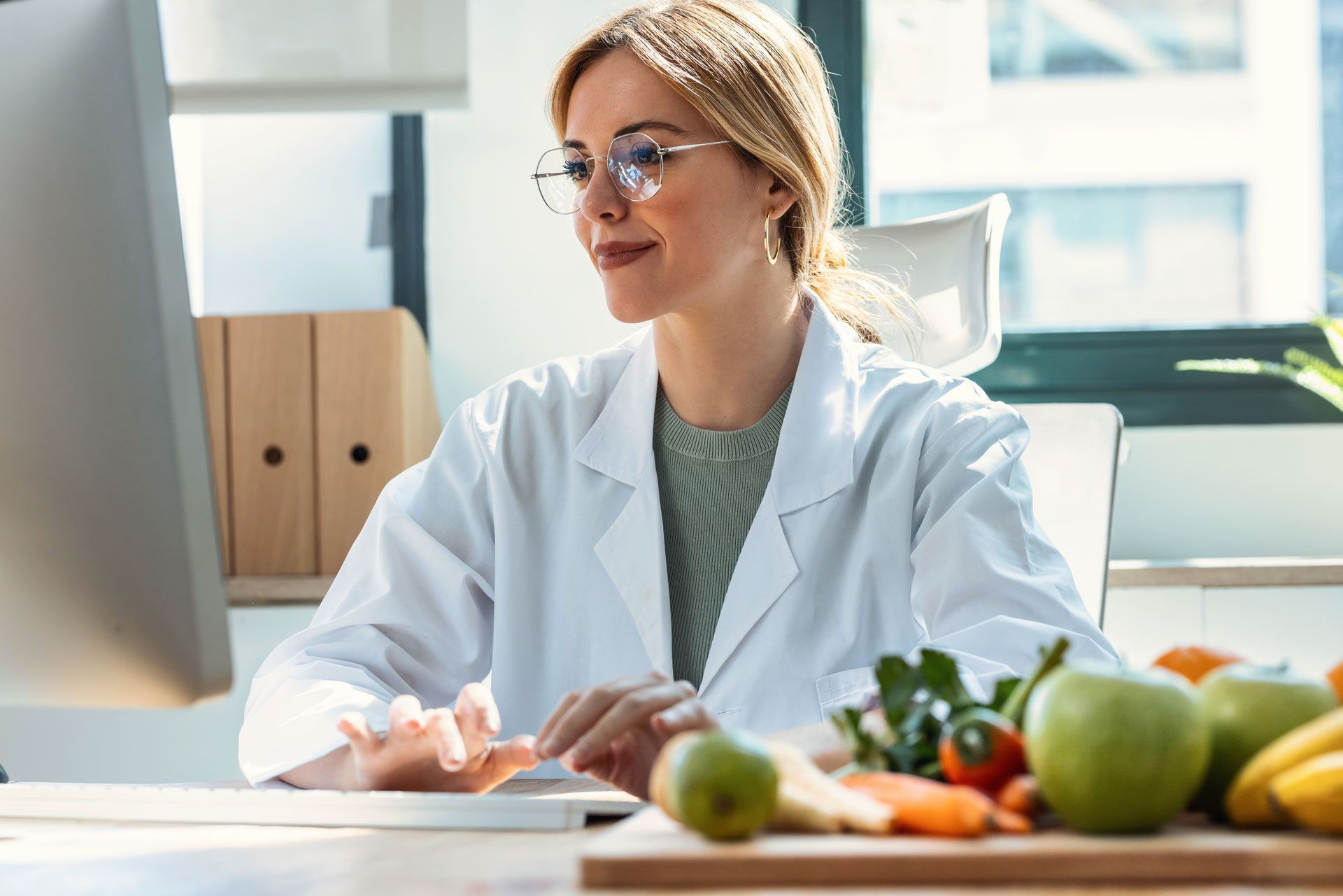 Dietitian in a white coat looking at a computer with fruit and vegetables on a cutting board in front of her.