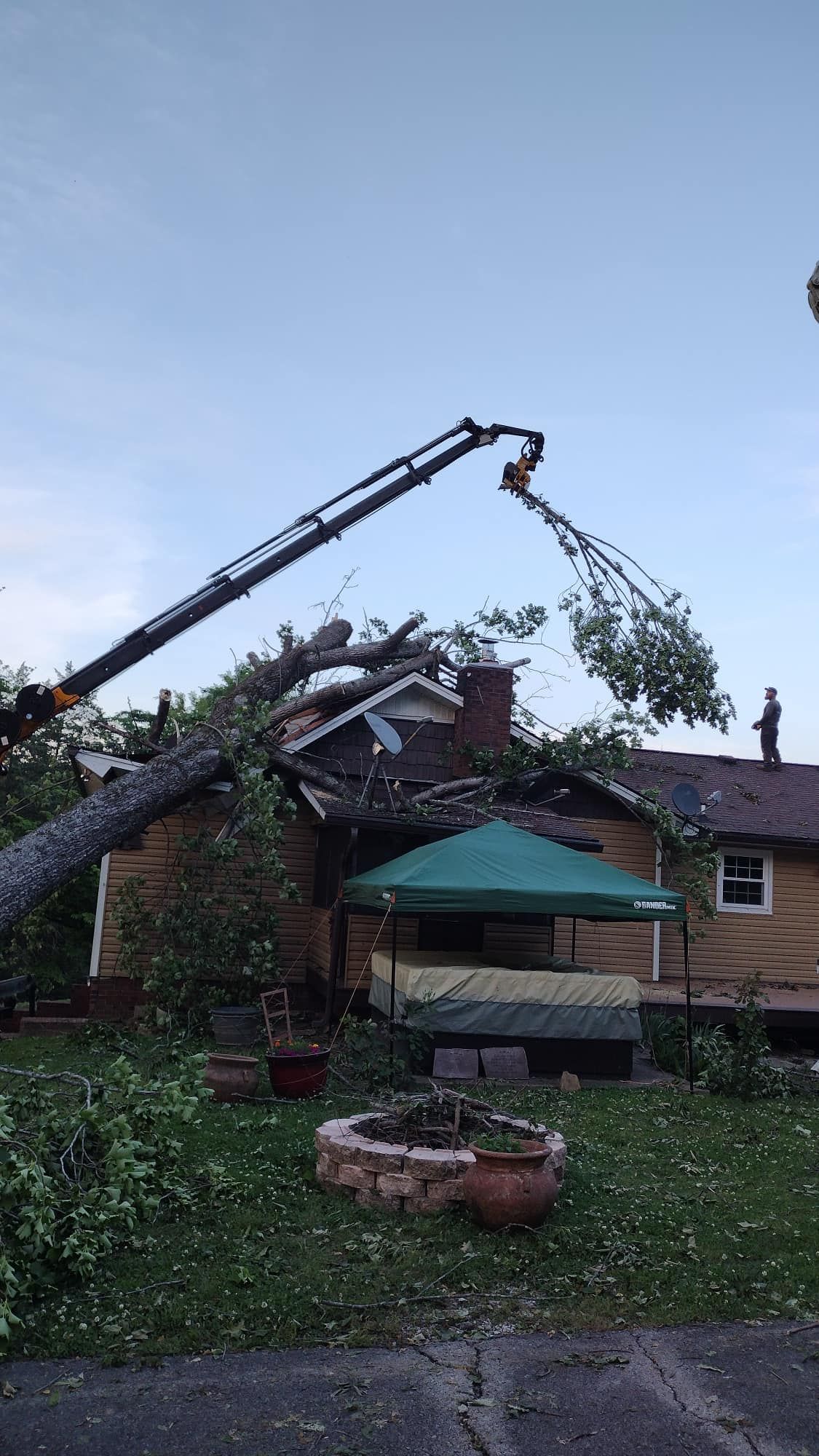 Crane lifting fallen tree from a backyard roof beside a house and patio umbrella
