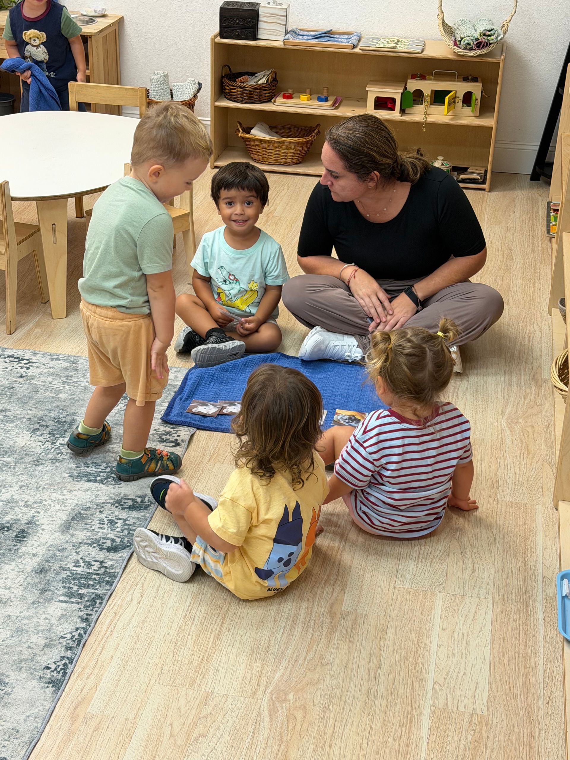 Children sit and work with an adult on a classroom rug near wooden shelves