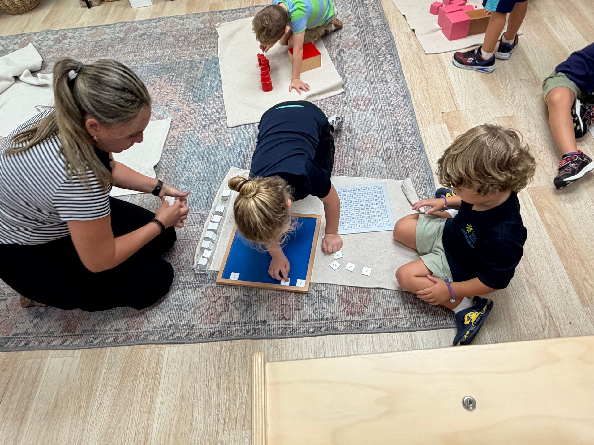 Children and an adult kneel on a rug, working on the hundred board in a Montessori classroom.