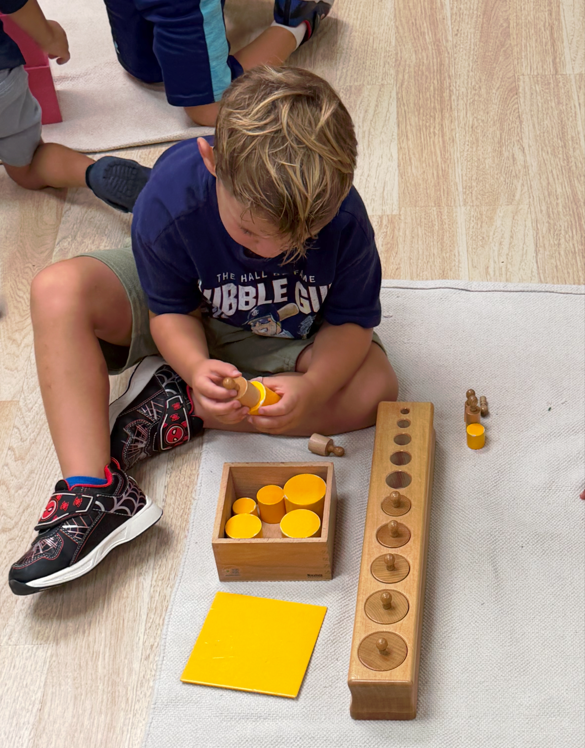 Child seated on the floor, working with the knobless cylinder and cylinder blocks in a classroom.