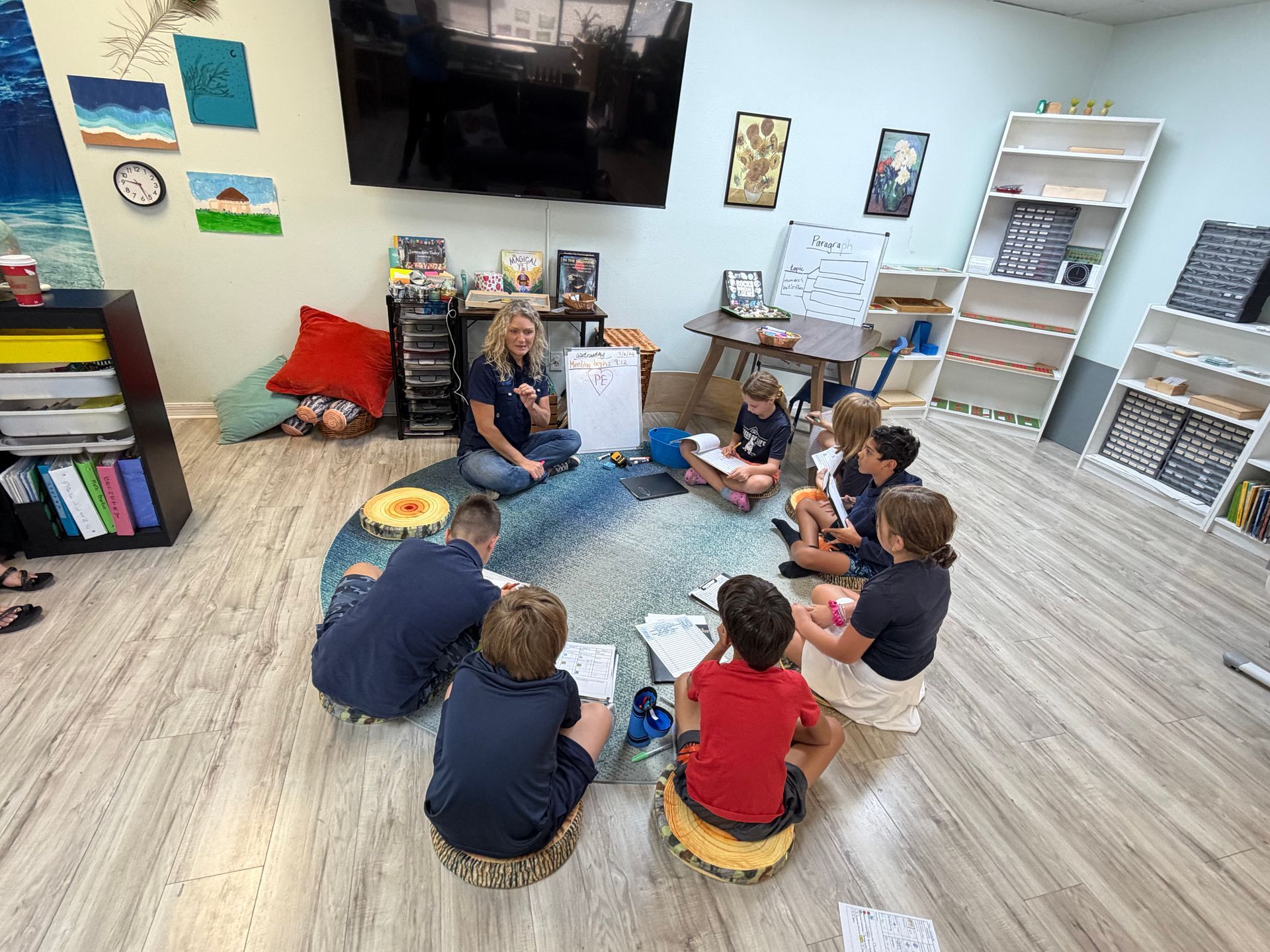 Children sit in a circle on a classroom rug with a teacher near a wall-mounted TV.