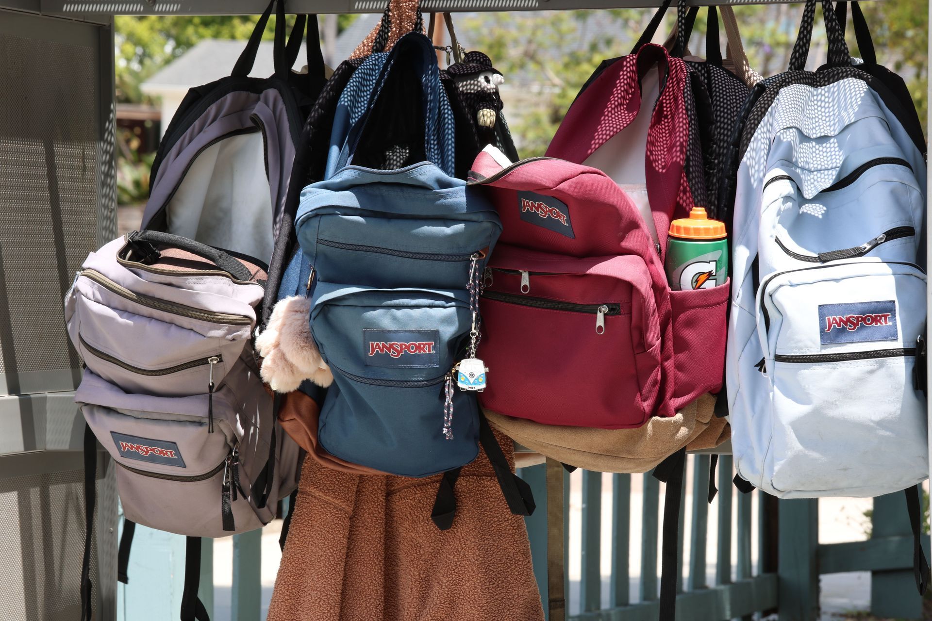 Backpacks hanging on a rack in various colors, including pink, teal, maroon, and light blue.