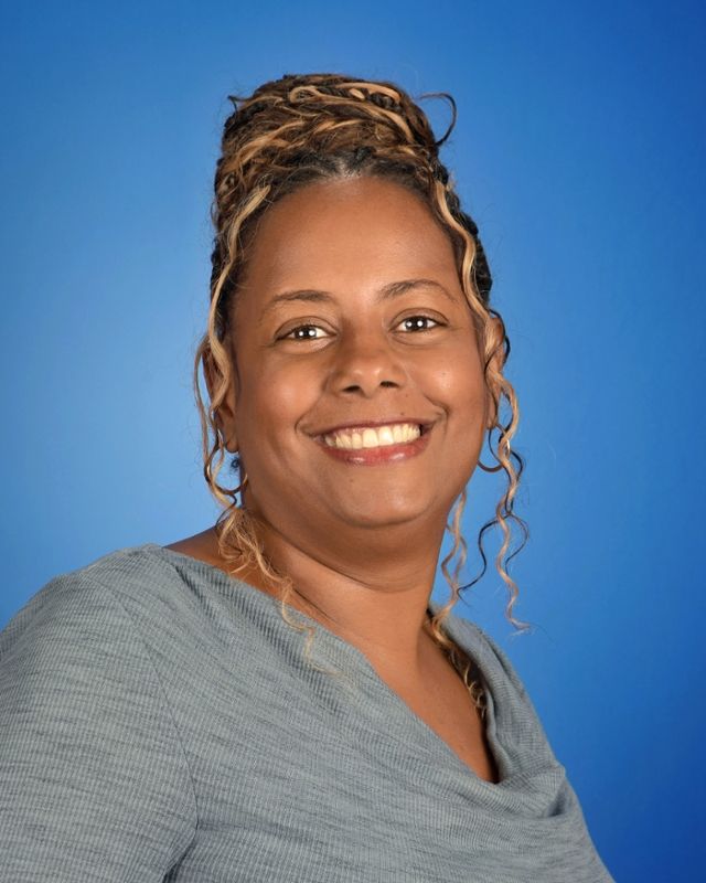 Smiling person with braided hair and gray top against a blue background