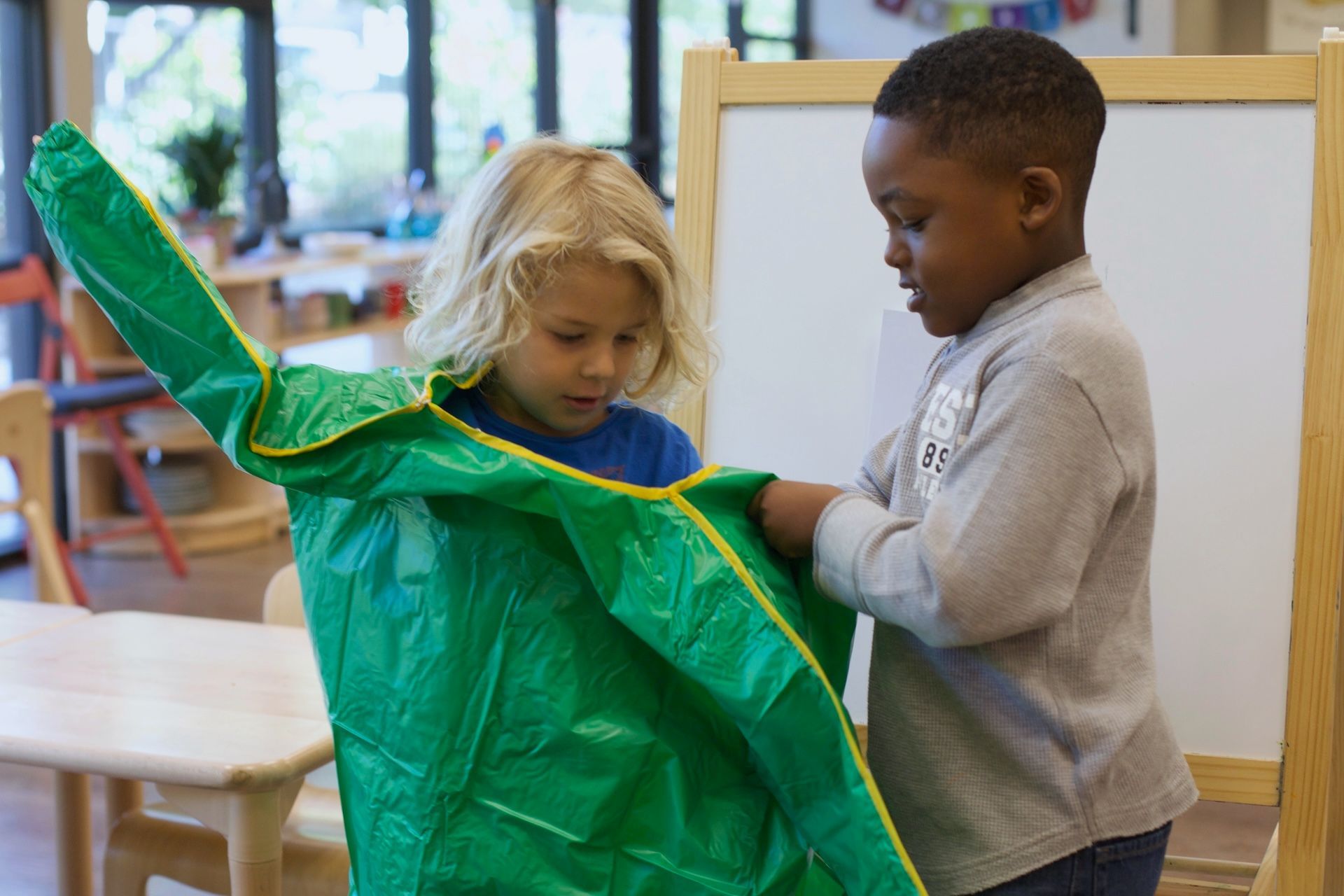 Two children in a classroom hold a large green costume or fabric sheet