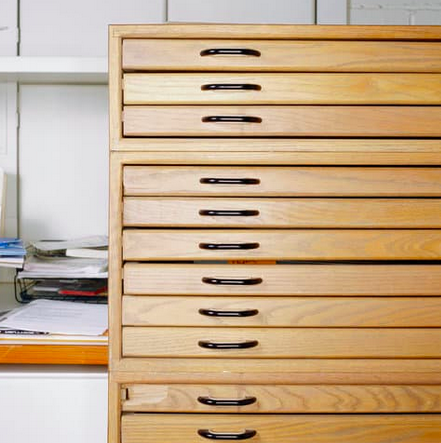 Wooden cabinet with multiple drawers, black handles. Partially visible cluttered shelf to the left.