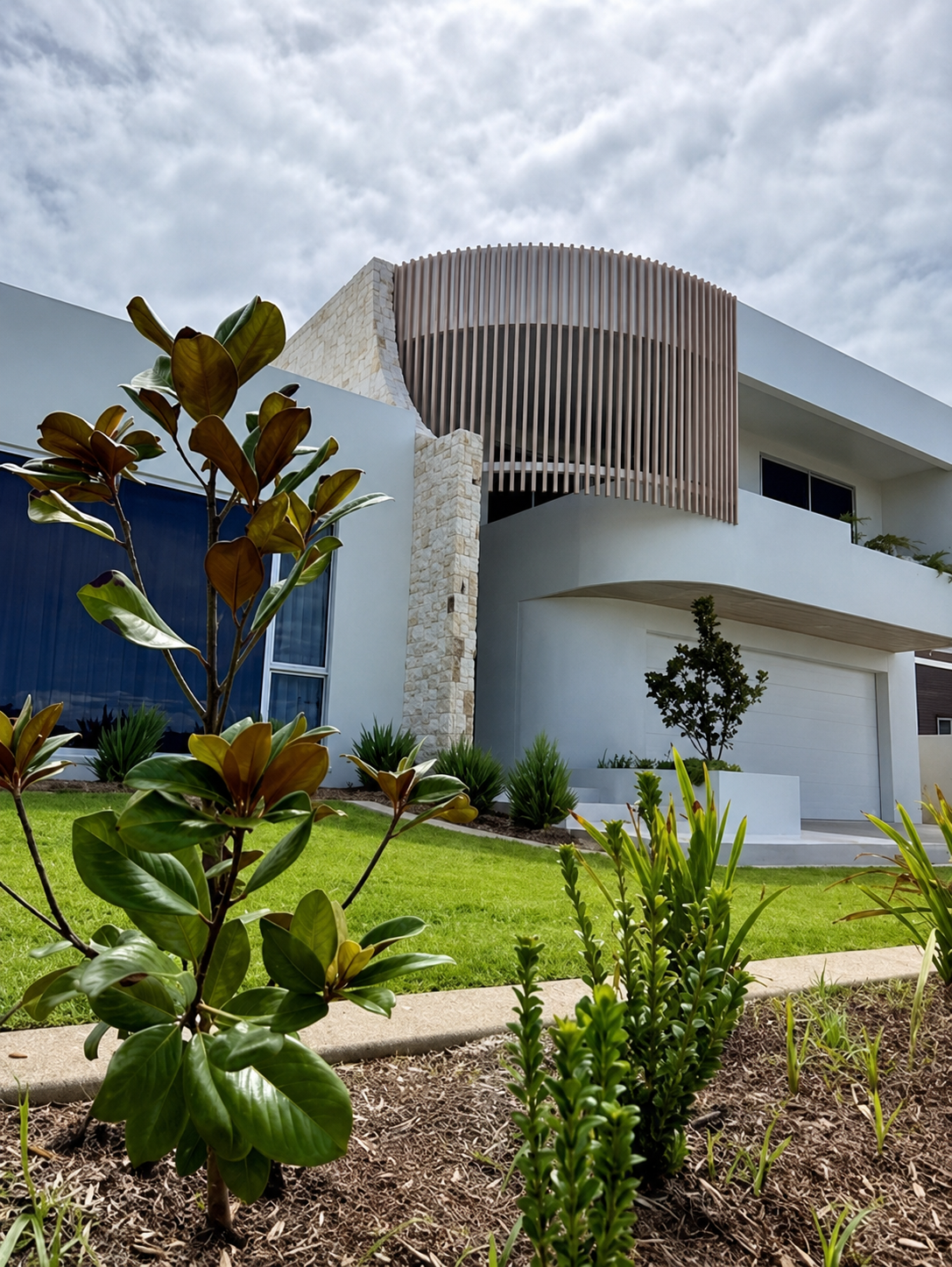 Modern white house with curved balcony, vertical slats, and landscaped green lawn with shrubs.