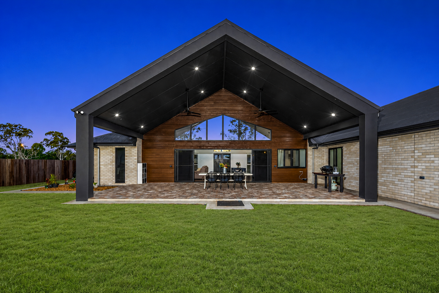 Modern house with a large covered patio, wood facade, and a wide green lawn at dusk