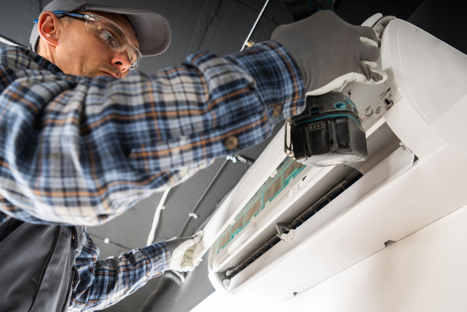 A technician in a plaid shirt installs an air conditioner with a drill while wearing safety glasses.