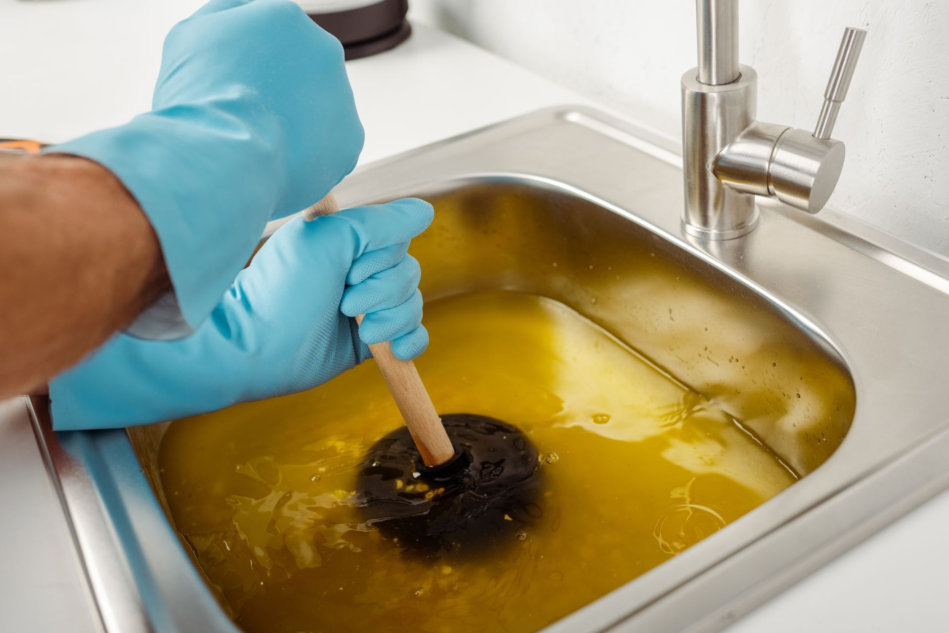 Person using a plunger in a sink full of yellow liquid, wearing blue gloves.