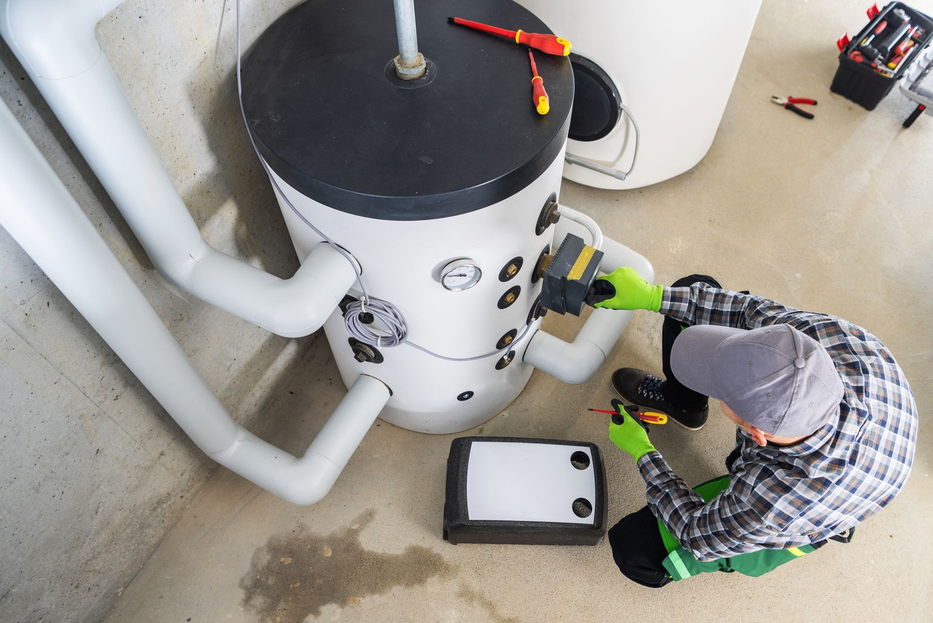 A person in green gloves works on a water heater. Tools and plumbing are visible in a basement setting.