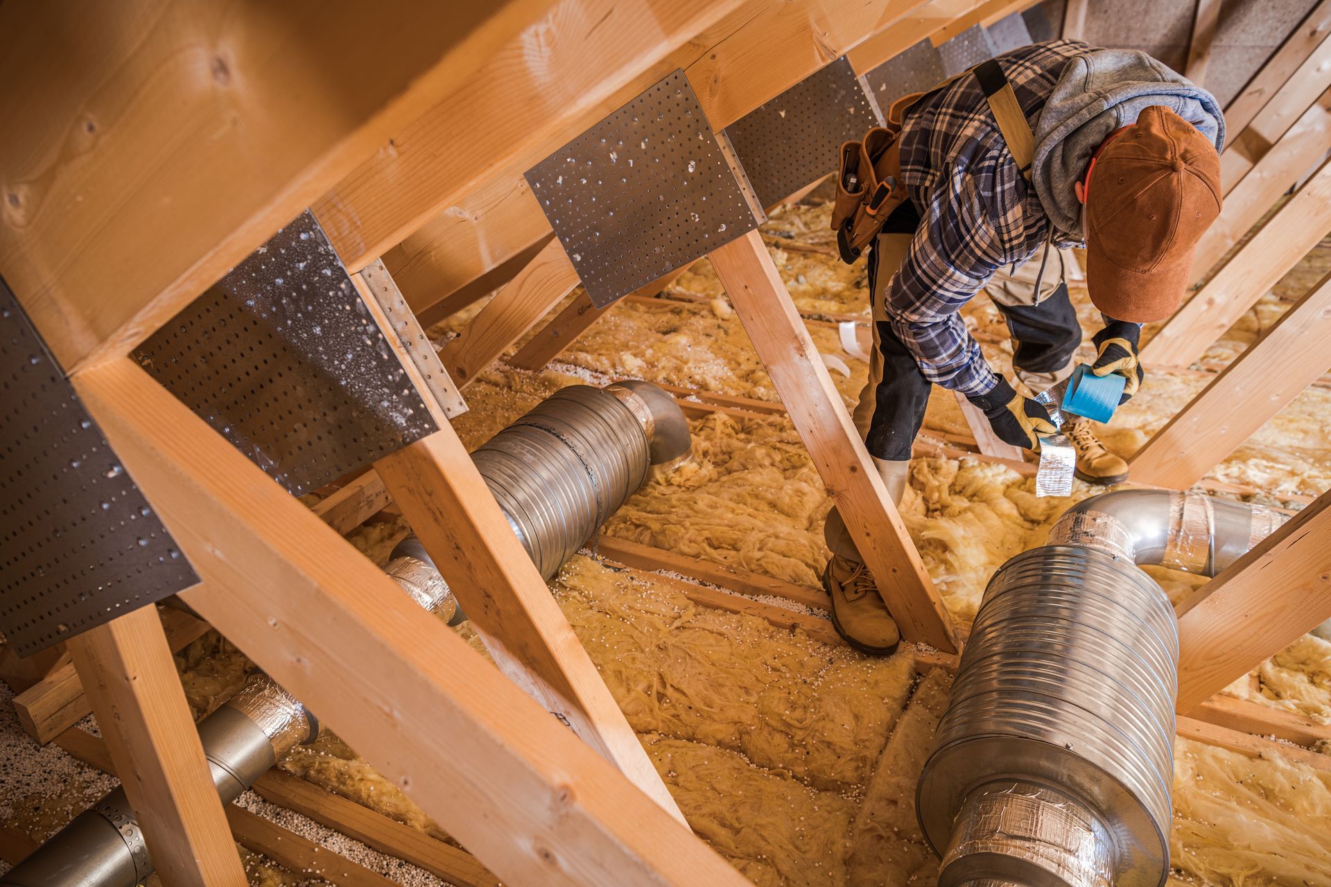 Person installing insulation in an attic with exposed wooden beams and ductwork.