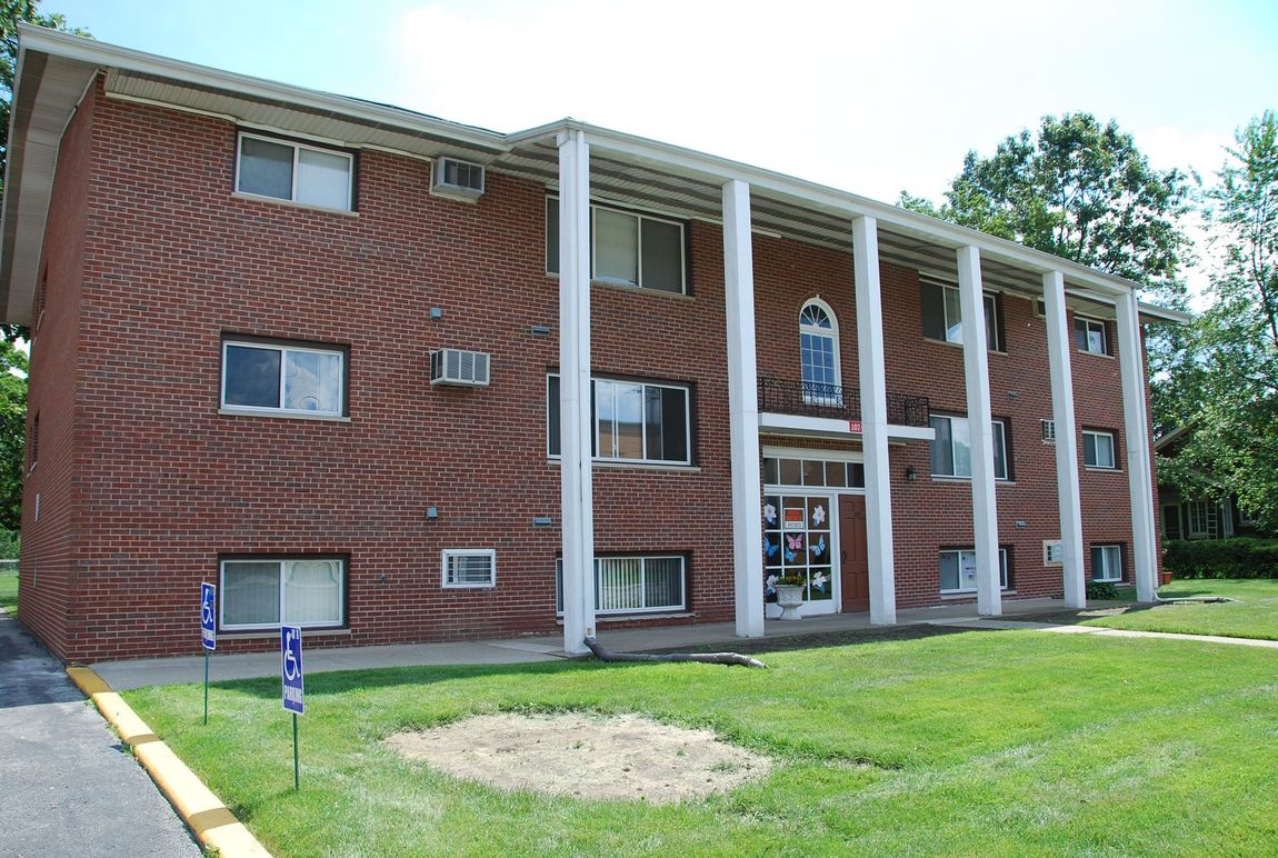 Two-story brick apartment building with white columns and a grassy lawn.
