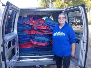 Woman in blue hoodie stands beside a van filled with folded red and blue blankets.