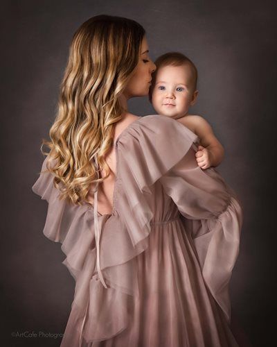 Woman in flowing pink dress kissing baby's head; gray background.