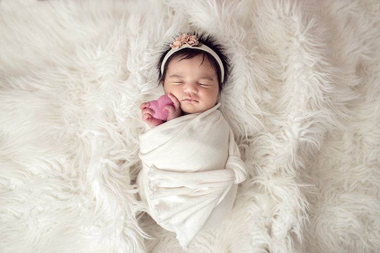 Newborn baby wrapped in white blanket, sleeping on fluffy white fur. Pink flower headband.