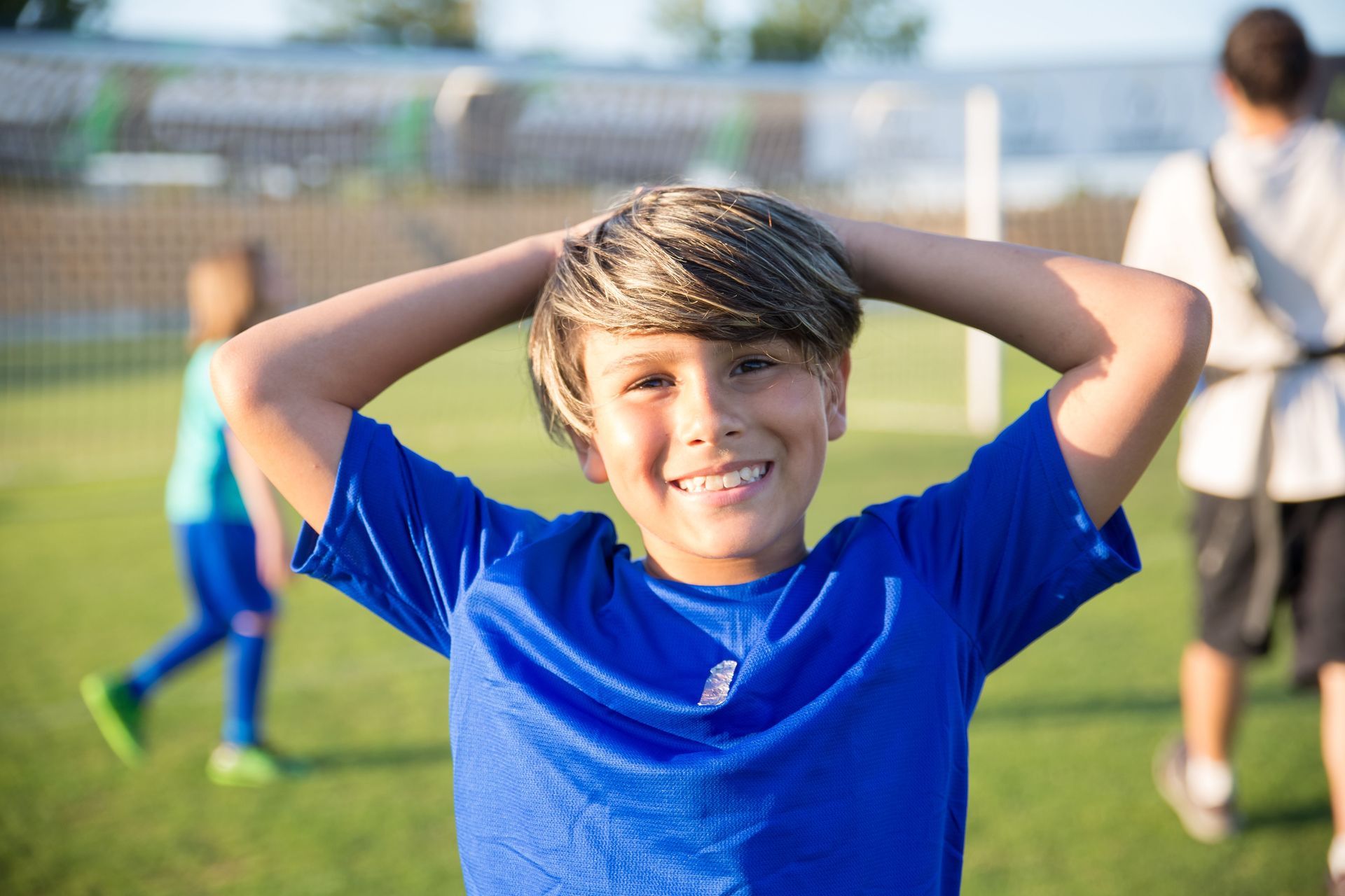 Boy in blue shirt smiling with hands behind his head on a soccer field, with other players in the background