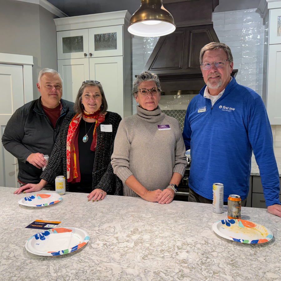 Four people standing behind a kitchen island with paper plates, a drink can, and a bright hanging lamp.