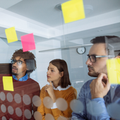 Team collaborating in an office, looking at notes on a glass wall with sticky notes.