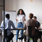 Small group meeting around a table in a modern office, with one person standing and presenting.