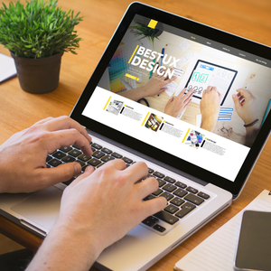Hands typing on a laptop displaying a web design site on a wooden desk рядом with a potted plant and notebook
