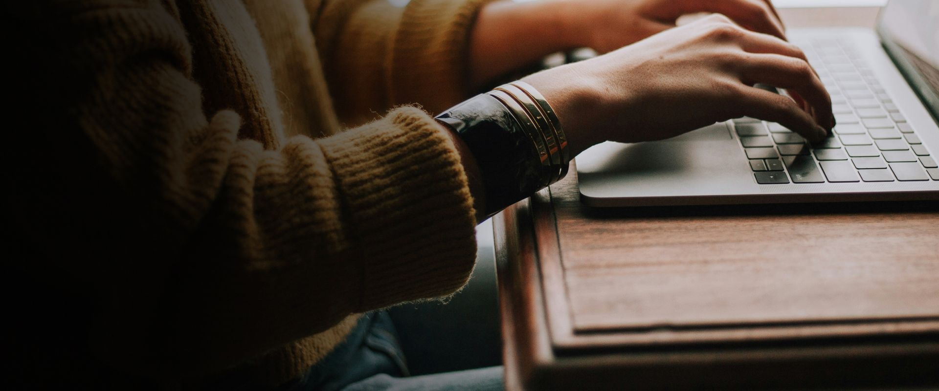Person typing on a laptop at a wooden desk, wearing a brown sweater.