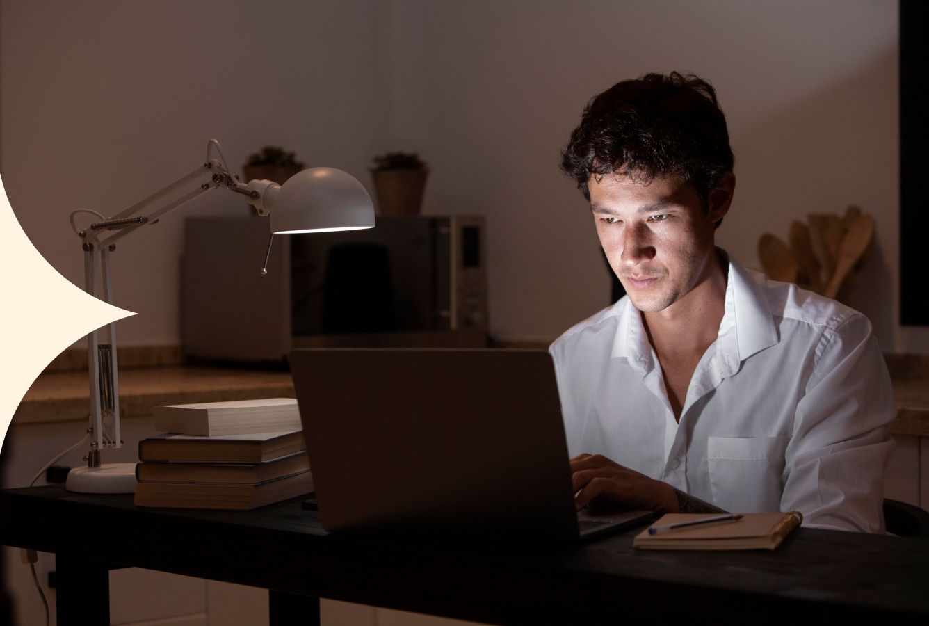 Person working at a laptop in a dimly lit room, illuminated by the screen beside a desk lamp.