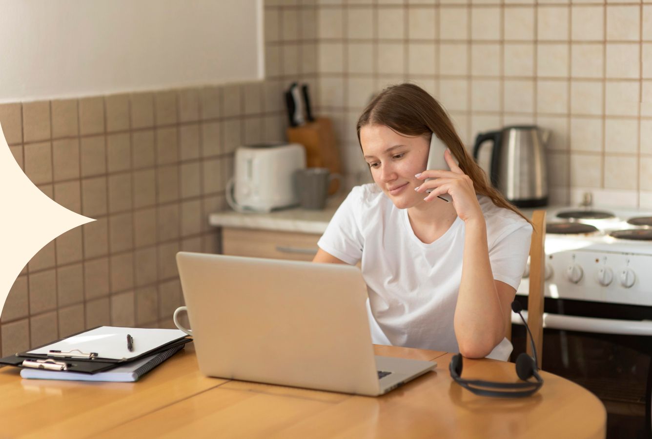Person working on a laptop at a kitchen table with a notebook, headphones, and a cup nearby