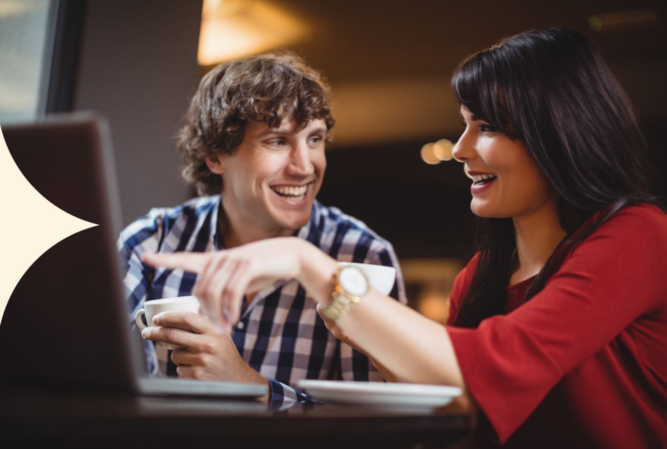 Two people smiling and talking at a laptop in a warmly lit café or office.