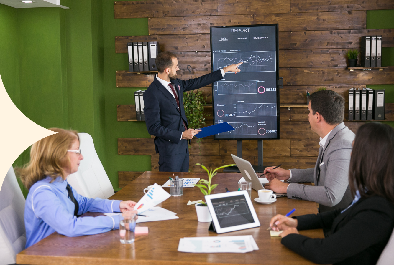 Business meeting in a wood-paneled office, with a presenter pointing at a chart on a screen and colleagues seated at a table.