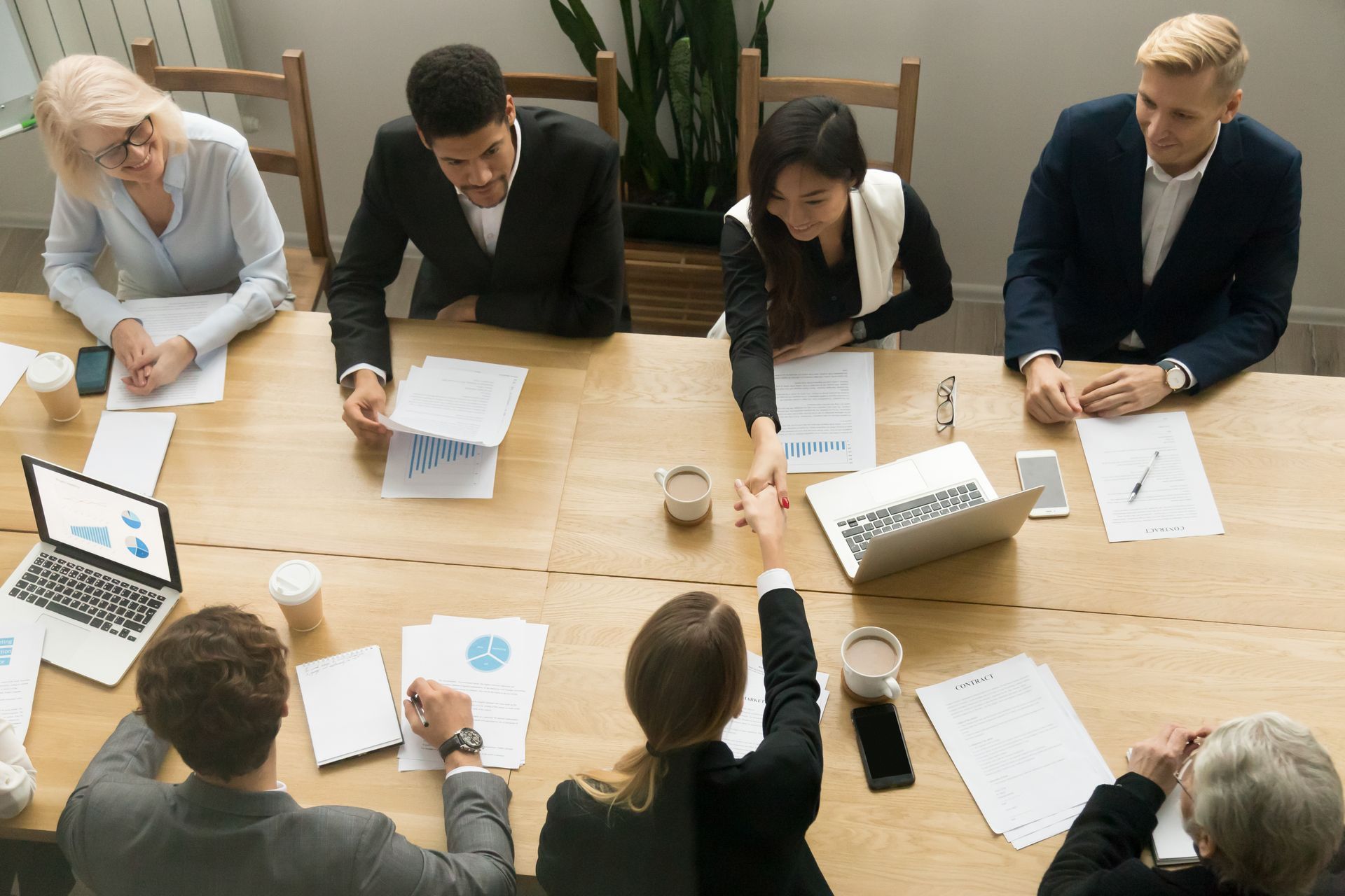 Team meeting around a wooden conference table, with laptops, papers, and charts in a bright office.