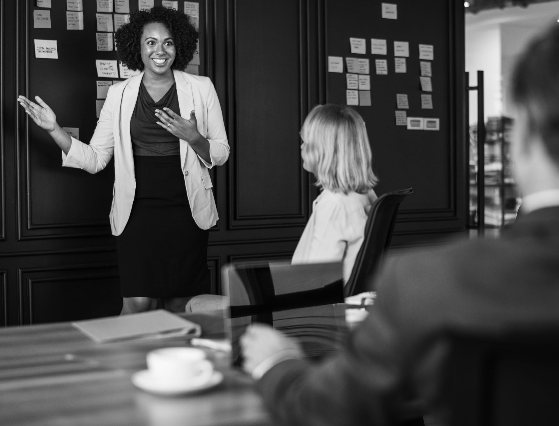 Presenter speaking to seated colleagues in a conference room, with a whiteboard and notes on the wall.
