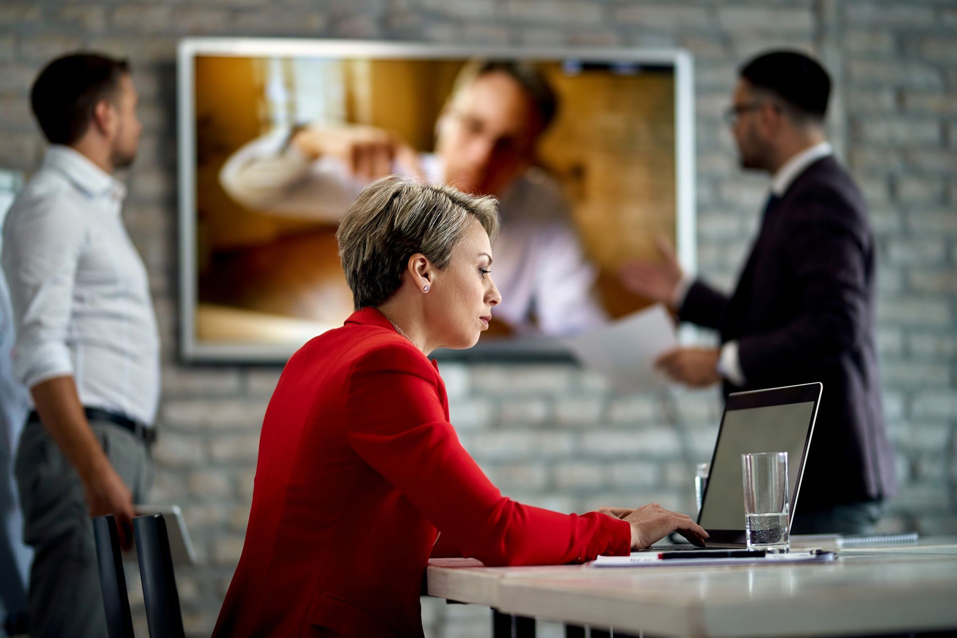 Meeting in office with three people, one seated at a laptop and two standing near a large screen.